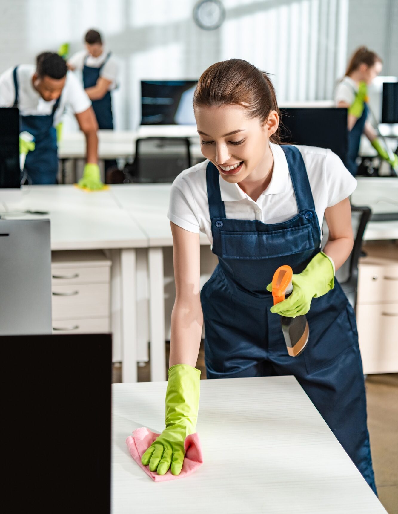 cheerful cleaner in overalls cleaning office desk with rag
